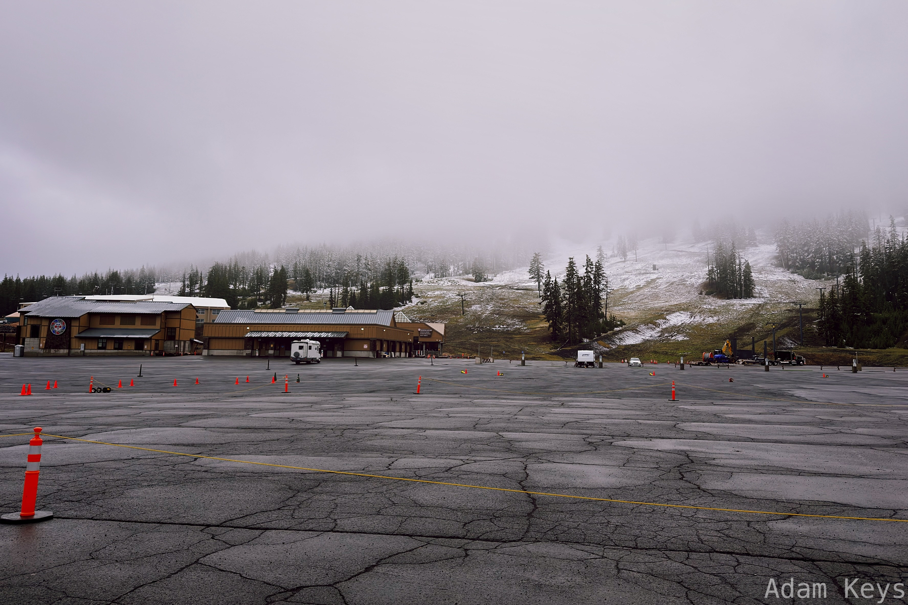 A large empty parking lot with orange traffic cones is situated in front of buildings at the base of a fog-covered mountain with sparse patches of snow and trees.